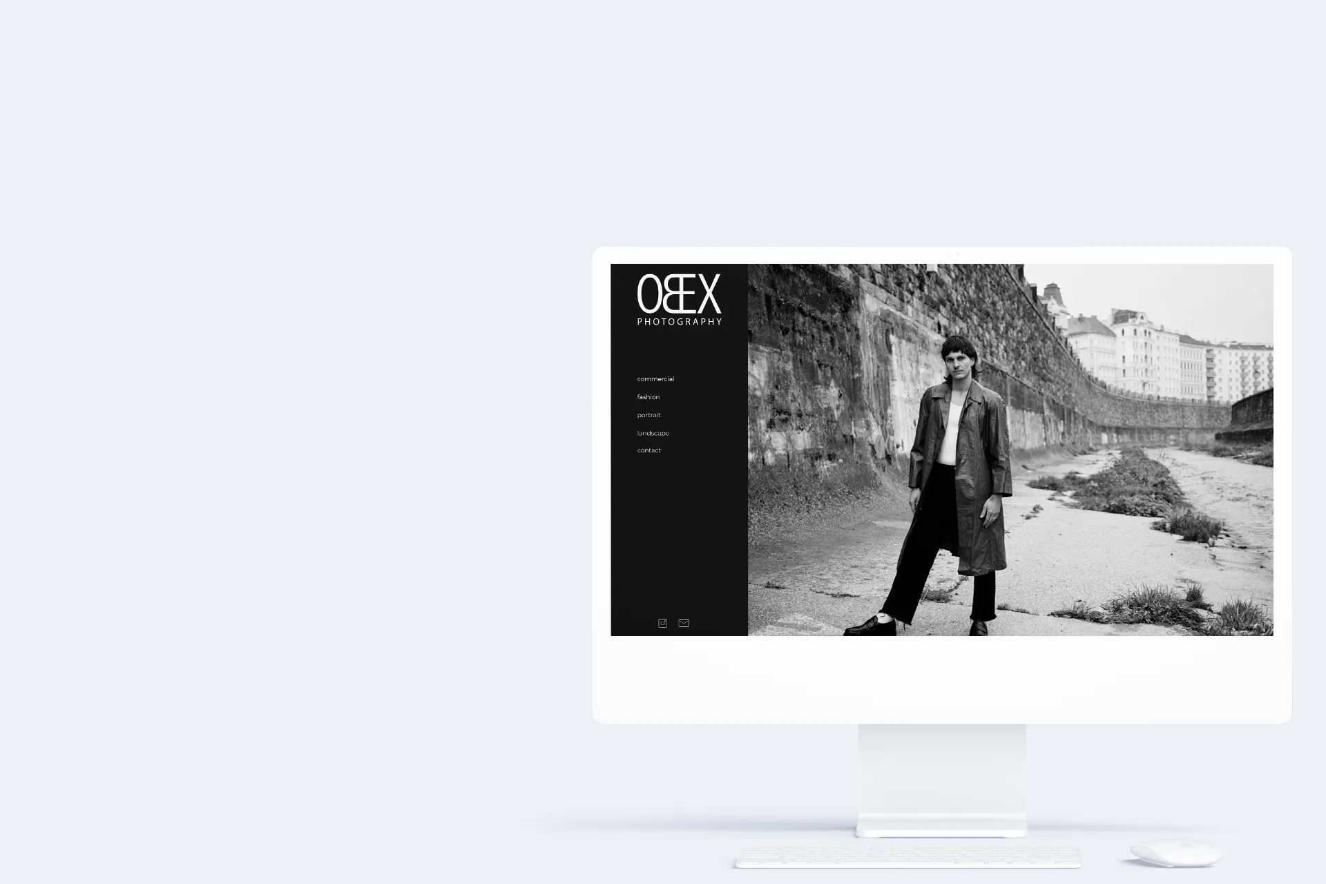 Ein weißer iMac. Auf dem Bildschirm ist die Startseite des Fotografen Obex Michael zu sehen, auf der ein Bild zu sehen ist, das eine Person in einem Trenchcoat mit einer markanten Frisur zeigt, die im Wien Kanal steht. 